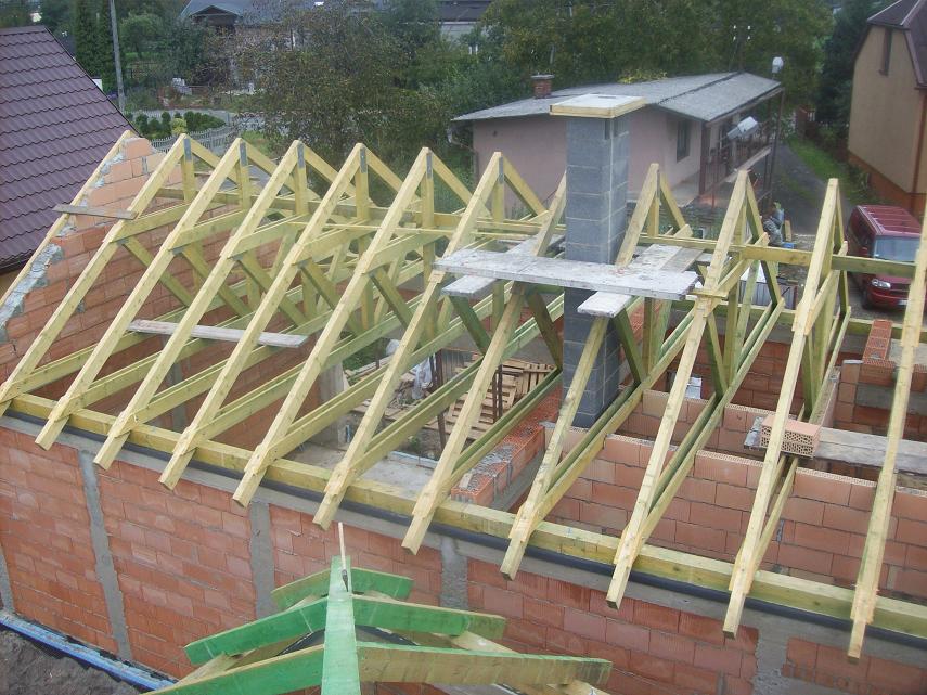 Wznoszenie konstrukcji dachu z drewnianych belek na murowanym budynku z cegły, z widocznym kominem w trakcie budowy i elementami deskowania.